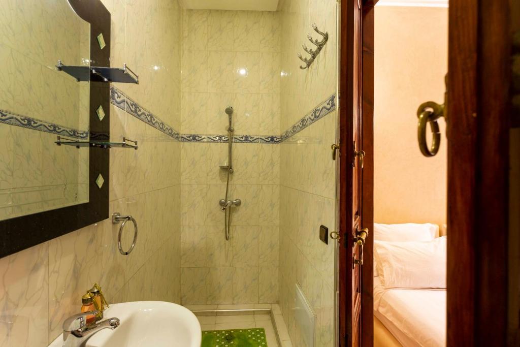 A small bathroom with cream-colored tiled walls, a wall-mounted shower, a sink with a mirror above it, and a towel ring. A wooden door on the right leads to a bedroom with a bed visible. A green bath mat is on the floor.