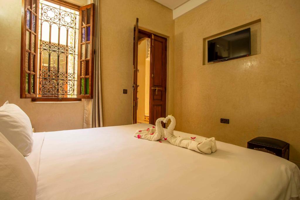 A cozy bedroom featuring a neatly made bed with white linens and towels folded into swan shapes, adorned with pink petals. The room has a window with decorative ironwork, allowing natural light to filter in. A flat-screen TV is mounted on the wall.