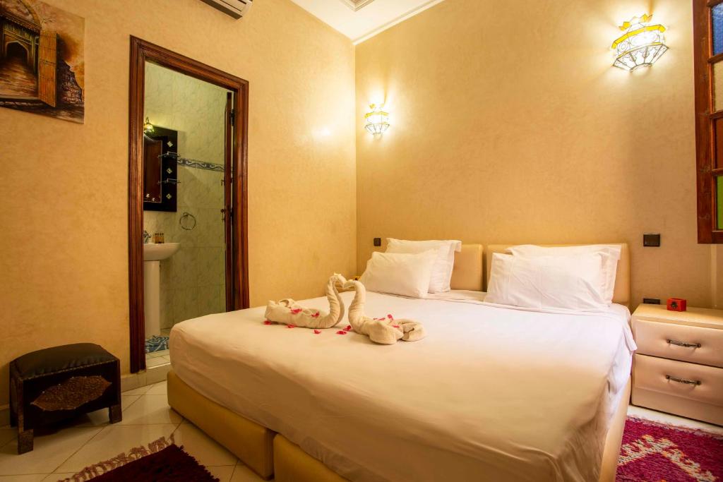 A cozy bedroom with a neatly made double bed, featuring white linens and decorative towel swans with petals. Warmly lit with wall sconces, the room has a small nightstand and an open door leading to a bathroom.