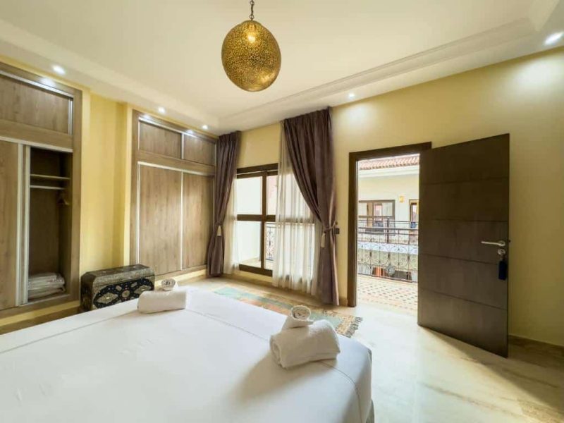 A bright, cozy bedroom featuring a large bed with white linens and rolled towels. The room has wooden closets, a patterned ottoman, and curtains on large windows. An open door leads to a balcony with a view of neighboring buildings.