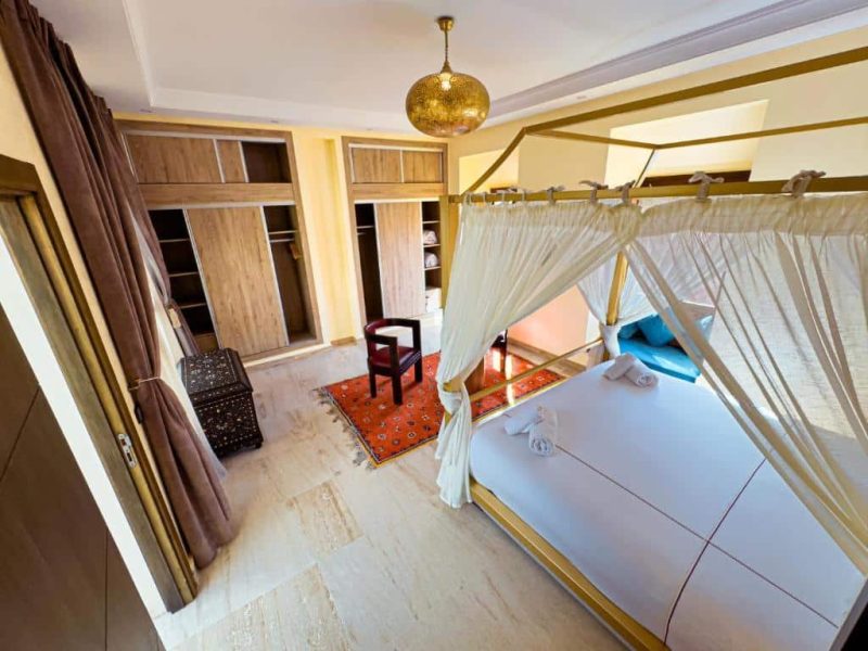 Bedroom with a canopy bed draped in white curtains. The room has wooden wardrobes, a red patterned rug, and a small chair. A gold pendant light hangs from the ceiling. Natural light streams in through a partially visible window with brown curtains.
