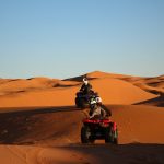 Two people ride ATVs over sand dunes in the Zagora Desert under a clear sky. The sun casts long shadows across the orange sand, highlighting the contours of the dunes.
