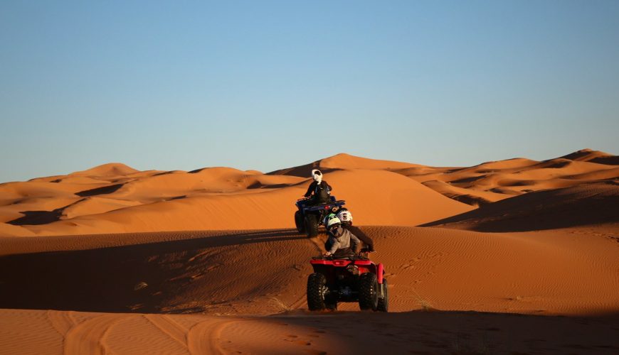 Two people ride ATVs over sand dunes in the Zagora Desert under a clear sky. The sun casts long shadows across the orange sand, highlighting the contours of the dunes.