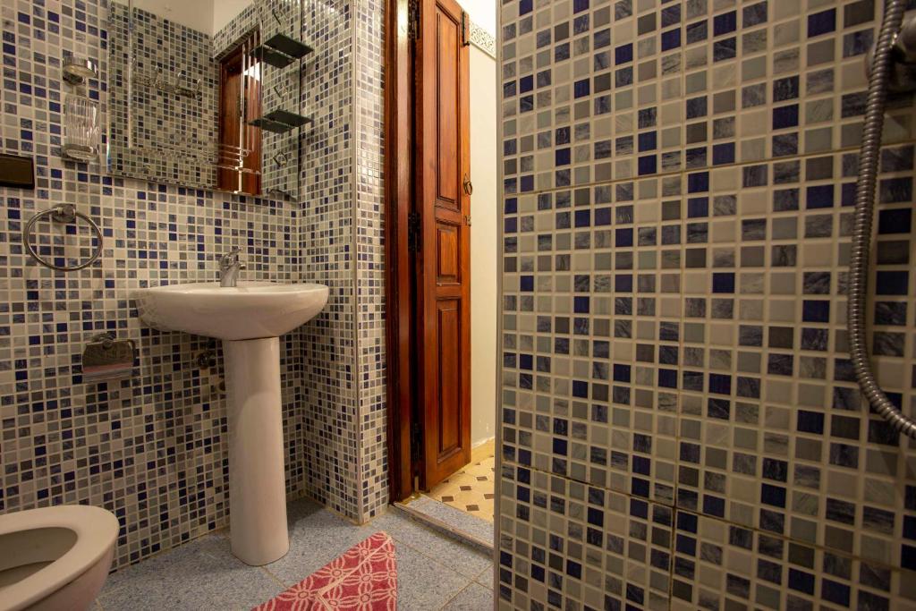 A bathroom with blue and white mosaic tile walls. It features a pedestal sink, a wall-mounted towel ring, and a wooden door. Theres a glimpse of a shower with a chrome fixture and a small section of a red and white patterned floor mat.