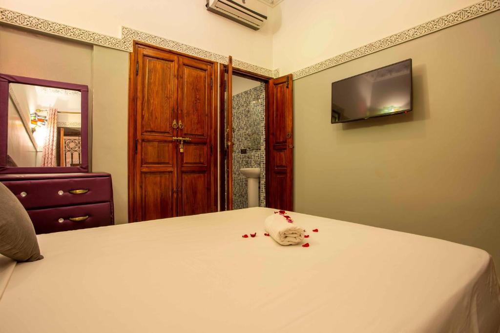 A cozy bedroom with a neatly made bed. Rolled towels and rose petals are on the bed. The room features wooden doors, a wall-mounted TV, and a dresser with a mirror. The bathroom door is slightly open, revealing mosaic tiles.