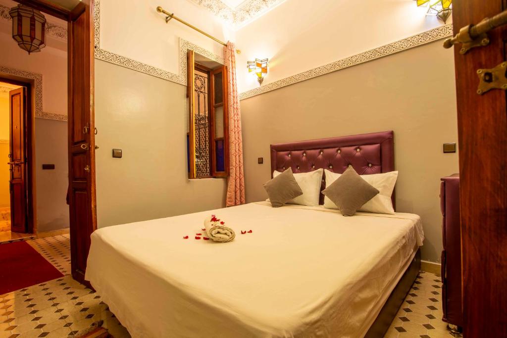 A cozy bedroom with dim lighting features a bed with a purple headboard, white linens adorned with two gray pillows, and a towel with rose petals. The room has patterned tile flooring and a window with curtains. A wooden door is partially open.