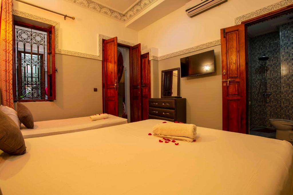 A cozy bedroom with a large bed adorned with a rolled towel and rose petals. The room has two wooden doors, a window with iron bars, a flat-screen TV, a dresser, and an open door leading to a tiled bathroom. Warm lighting creates a welcoming atmosphere.