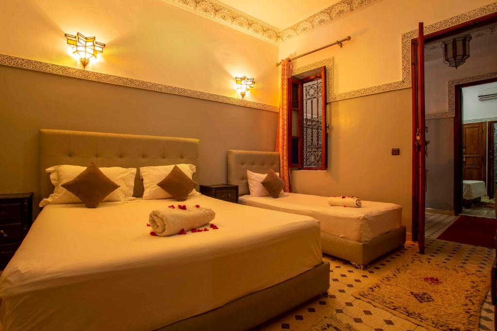 A cozy bedroom with warm lighting features a double bed and a single bed, both with white linens and brown accent pillows. Decorative floor tiles and wall patterns add elegance. A window with drapes is partially open, and towels are neatly placed on the beds.