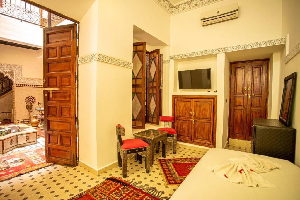 A cozy room with traditional decor featuring wooden doors and furniture. The room includes a TV, a small table with two chairs, decorative rugs, and patterned tile flooring. An adjoining area with a decorative staircase is visible through the open door.