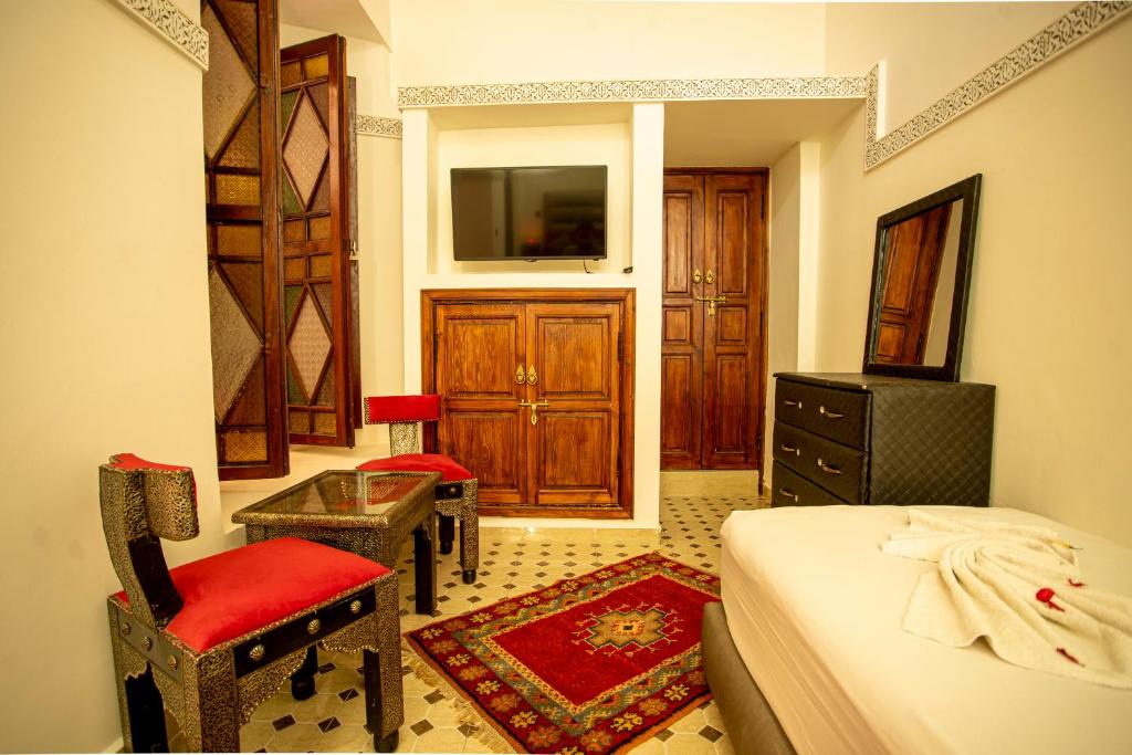A cozy room with traditional decor, featuring a wooden cabinet and TV, red cushioned chairs, a small table, a patterned rug, and a bed with folded towels shaped into swans. Walls have intricate designs and natural light filters through a window.