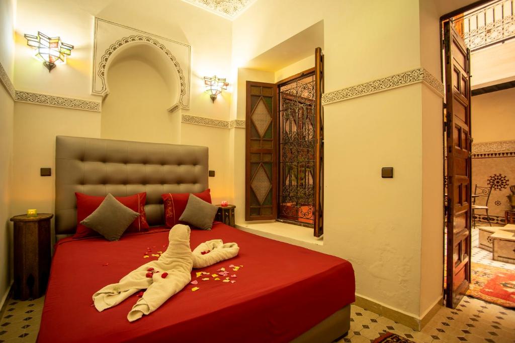 A cozy bedroom with an ornate ceiling and traditional decor. The bed has a red coverlet with a towel sculpted into a swan shape and scattered rose petals. Warm lighting and an open wooden window create a welcoming atmosphere.