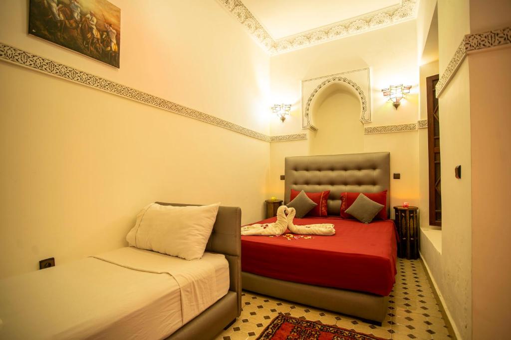 A cozy bedroom with a single bed and a double bed. The room features warm lighting, red bedding, decorative pillows, a patterned rug, and ornate wall details. A towel is folded into a swan on the double bed. A painting hangs above.