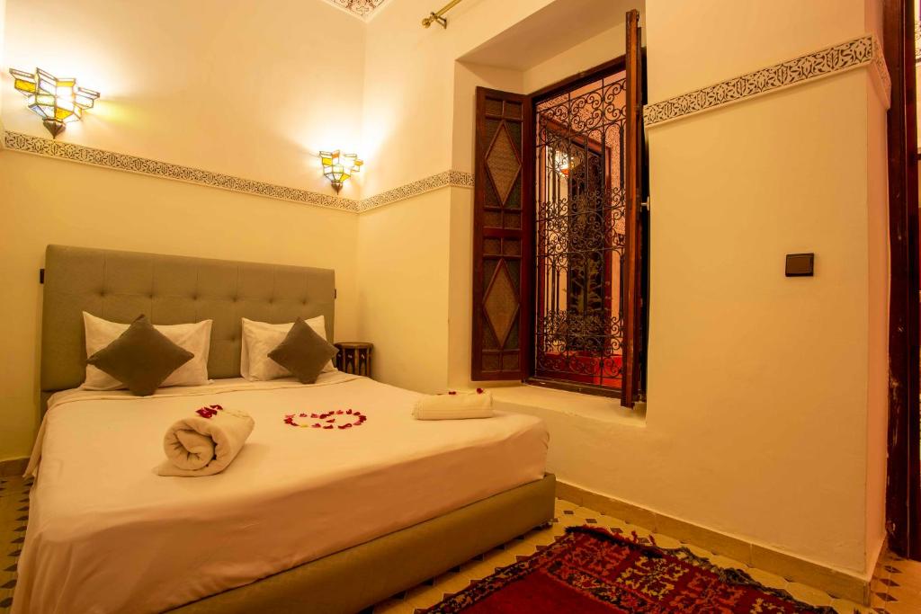 A cozy bedroom featuring a neatly made bed with white linens, three pillows, and rolled towels adorned with flower petals. Soft lighting comes from ornate lamps, and a window with decorative ironwork lets in warm light. A patterned rug lies on the floor.