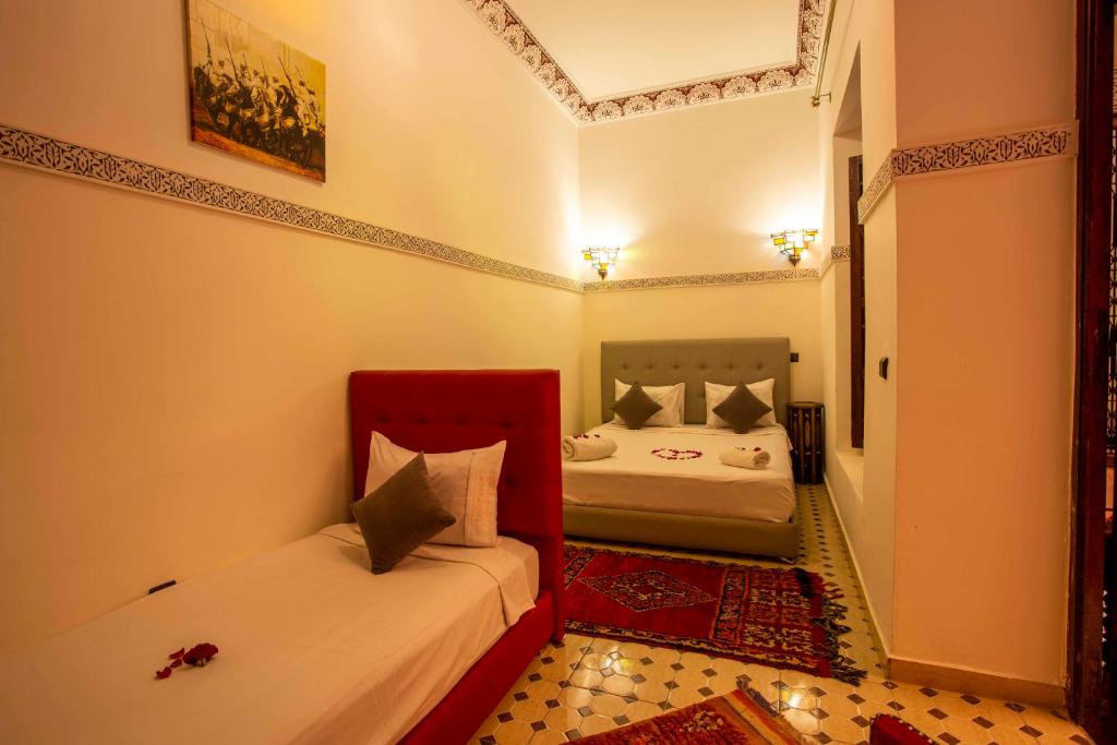 A cozy room with warm lighting features a single bed with a red headboard and a double bed with a gray headboard. Each bed is adorned with white and gray pillows. The floor has patterned tiles and red rugs. Wall decorations include a framed artwork.